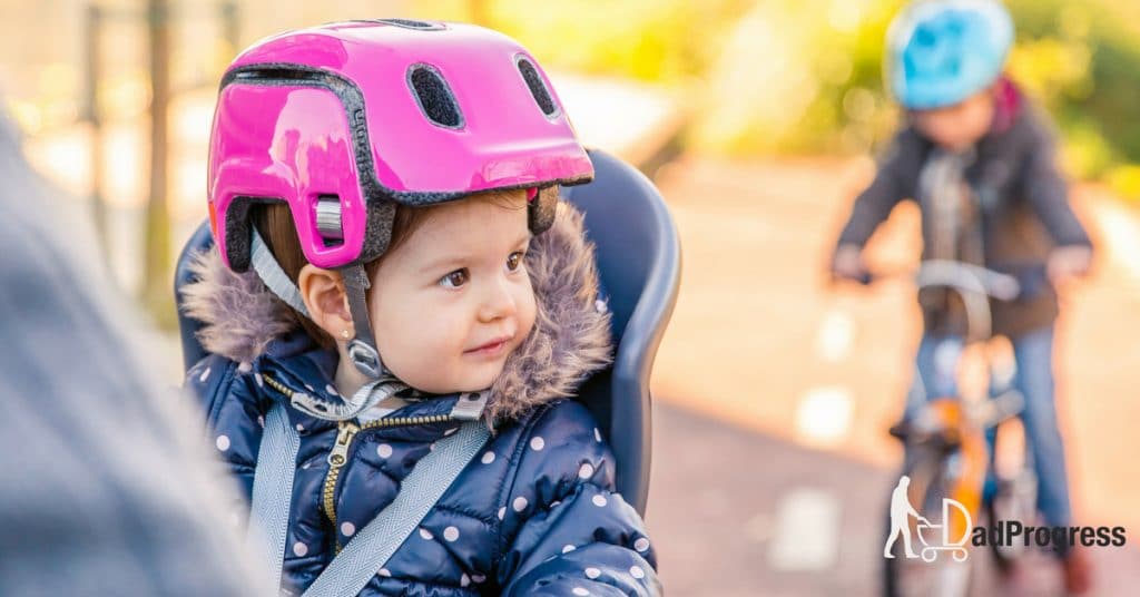 A toddler sitting on a bike chair and she has a helmet