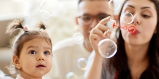 Mom blows bubbles for a young toddler, dad in the background