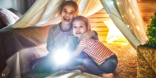 Two girls under a blanket and direct a flashlight directly at the viewer