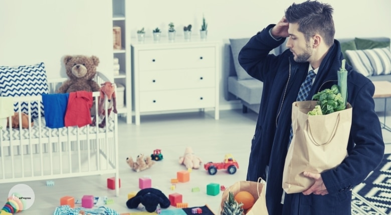 Man holds a grocery bag and looks the mess in the room- living room is full of toys and there's a regular crib on the left