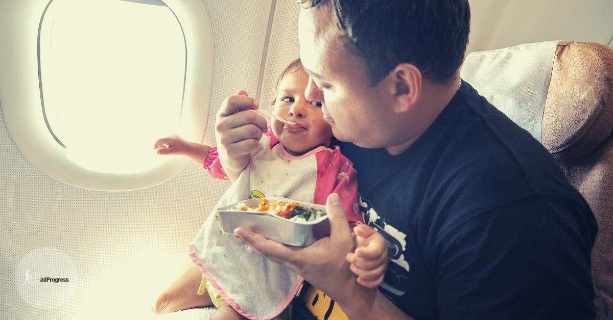 Father sitting in an airplane seat and feeding a toddler who's sitting on his lap