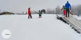A toddler with skiis on snow and an instructor before him. On the right there are two other people going up the small mount