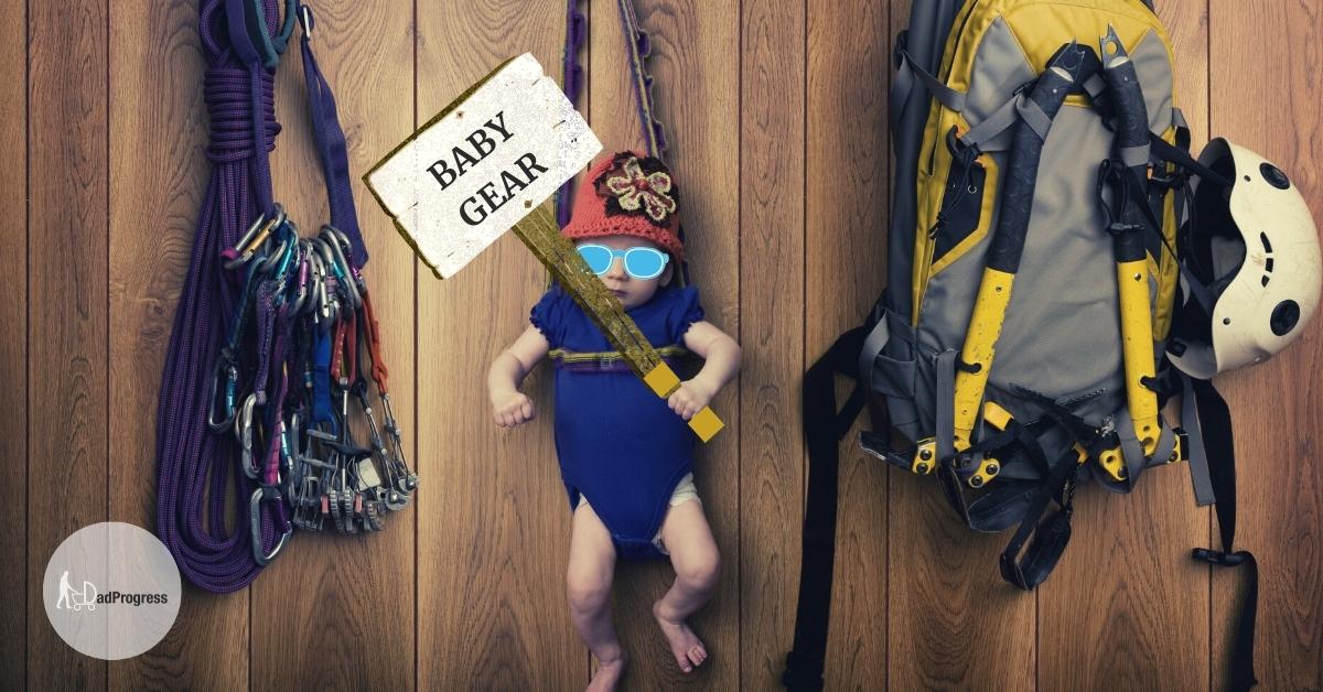 Baby stuff for dads featured image- mountaineering equipment hanging on the wall and also an infant in a baby carrier, he’s holding a sign “Baby Gear” in his left hand and wears blue sunglasses