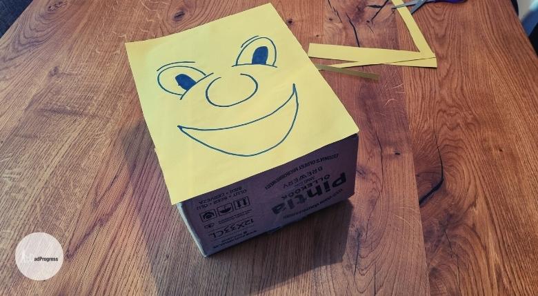 A yellow paper is glued on a cardboard box and there's a smiling face drawn on it