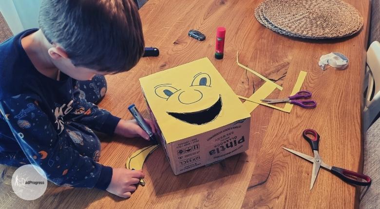A yellow paper is glued on a cardboard box and there's a smiling face drawn on it, mouth is now open. A toddler is drawing something on a table on the left of the carboard box