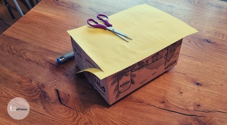 Yellow paper is glued on a cardboard box and the box is on a wooden table. Scissors are seen on the box, they are used to cut the corners of the paper